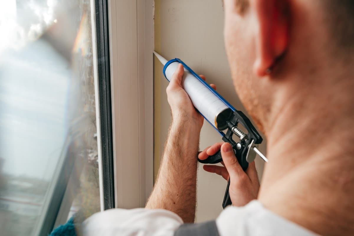 Young man wearing overalls sealing cracks between window and trim