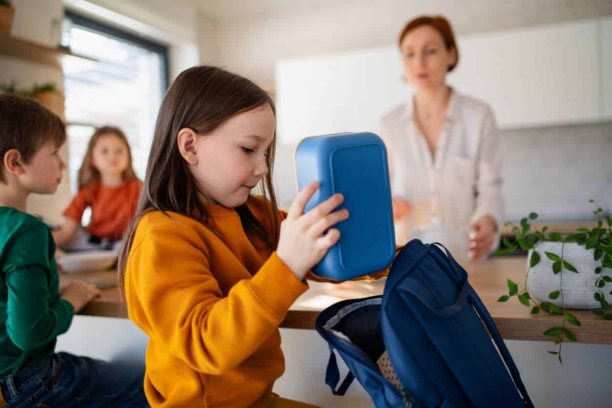 A child packs her lunchbox in her school backpack, with her mother and siblings in the background.