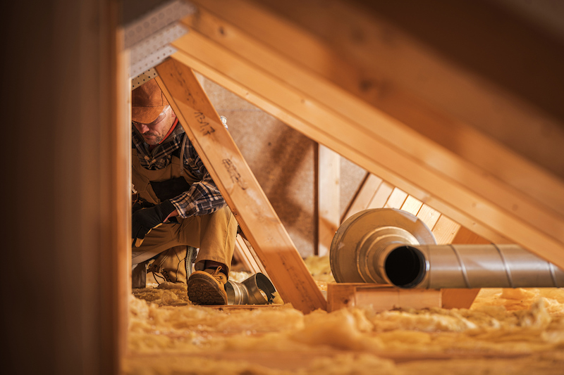 A contractor inspects the fiberglass insulation and vents inside an attic space.